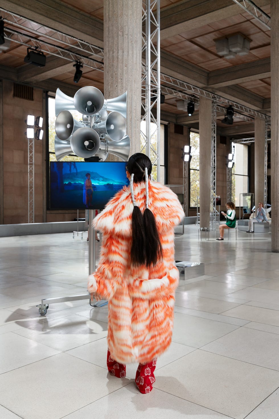 a person is standing in a modern gallery space, wearing a vibrant orange and white fur coat