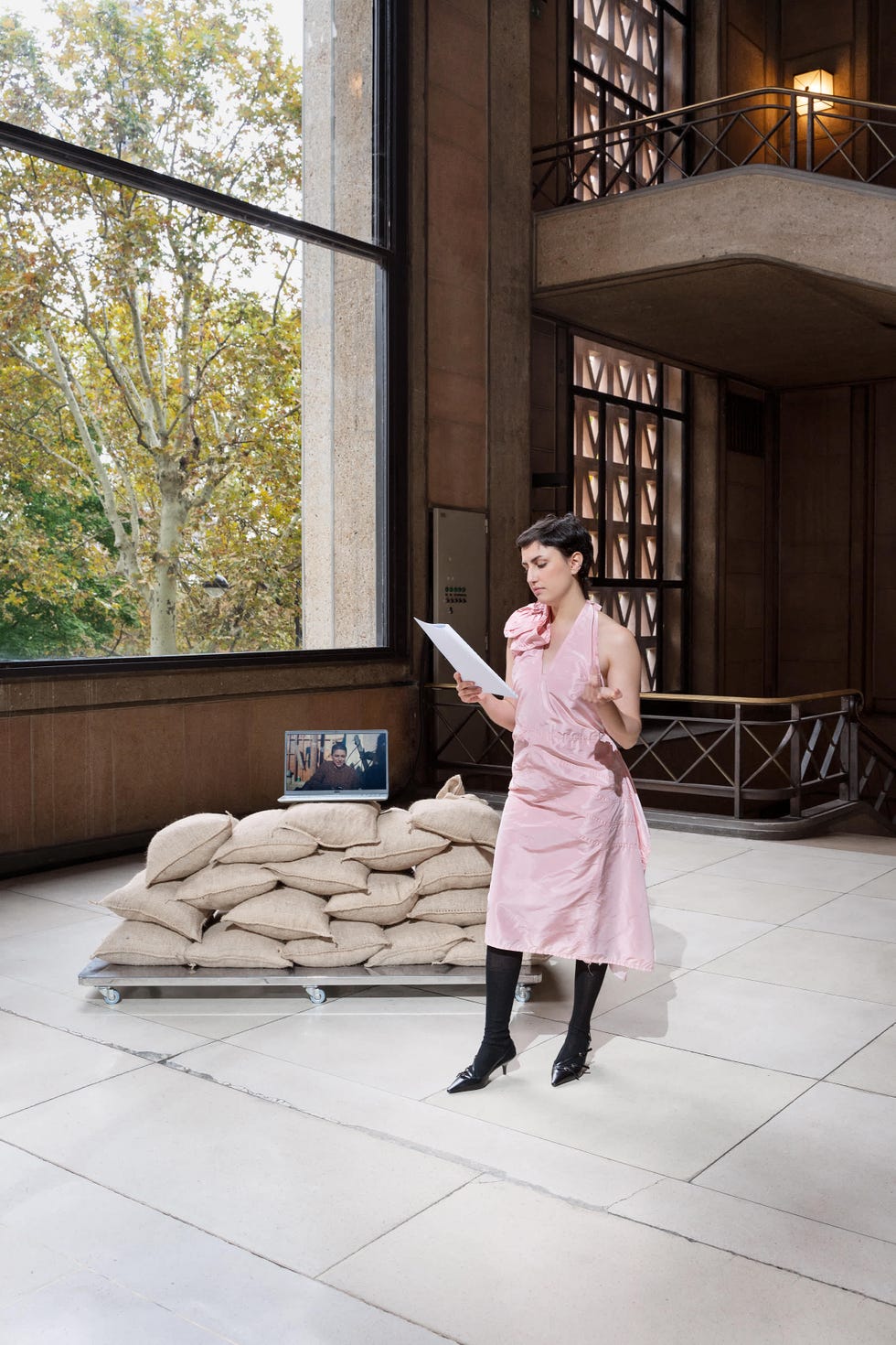 in a spacious indoor setting with large windows allowing natural light a stack of burlap bags is positioned on a rolling platform a person is standing in front of the bags, wearing a pink dress with a decorative element