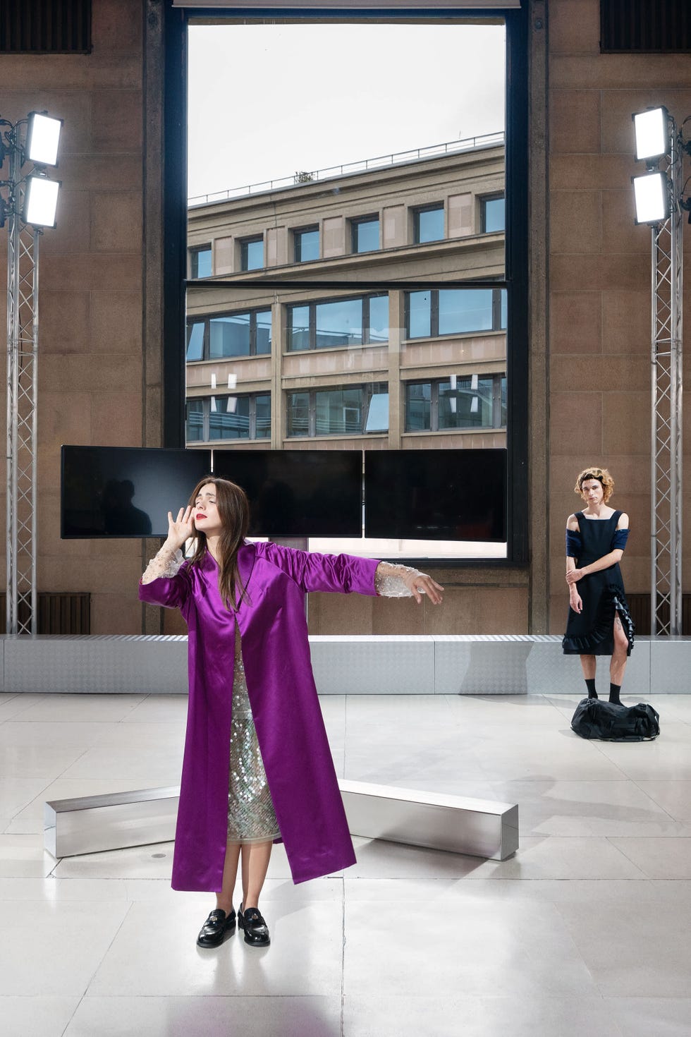 a scene featuring two figures on a minimalist stage with a large window displaying an urban building one figure is dressed in a long purple robe over a shiny silver dress striking a dramatic pose