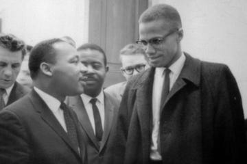 martin luther king jr and malcolm x speak to one another while standing in front of other men inside, king wears a suit jacket, collared shirt and tie, malcolm wears a coat, collared shirt, tie and glasses
