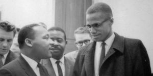 martin luther king jr and malcolm x speak to one another while standing in front of other men inside, king wears a suit jacket, collared shirt and tie, malcolm wears a coat, collared shirt, tie and glasses
