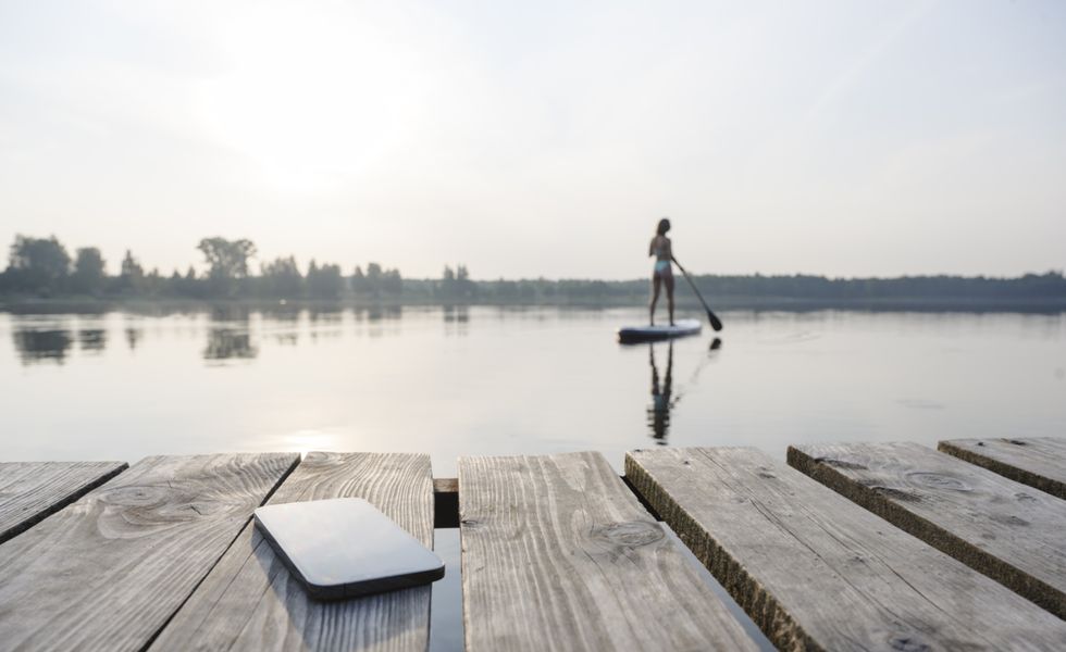 mobile phone on jetty with woman doing standup paddleboarding in background at sunset