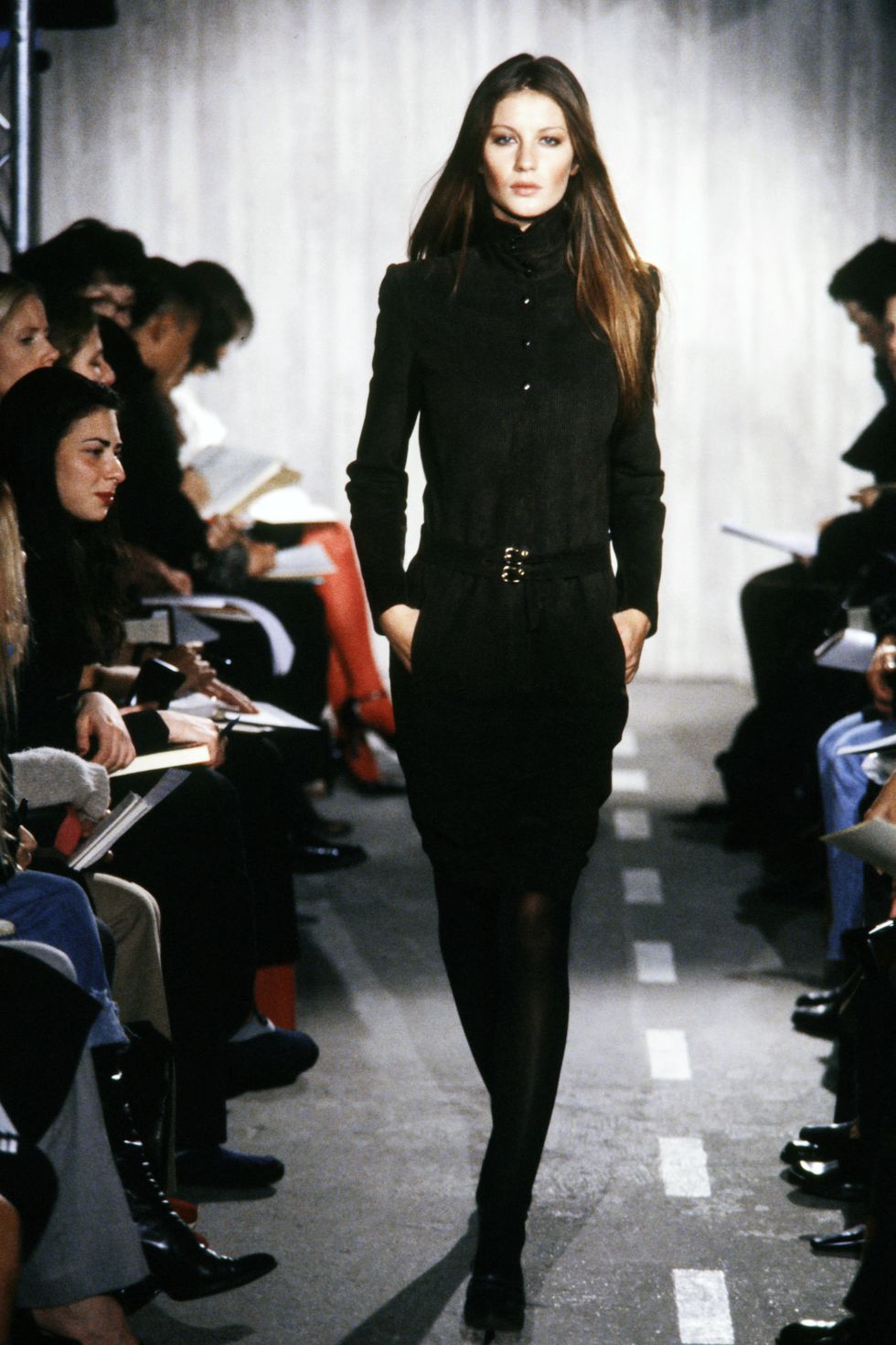 gisele bundchen walks with her hands in the pockets of her wall black outfit as people watch from seats on either side