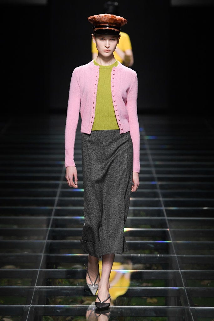 a model wearing a pink cardigan green knit top grey pencil skirt and light grey pointed toe slingback pumps with a brown leather paperboy cap on the prada rtw fall 2024 runway in a roundup of the best cardigan outfits 2024