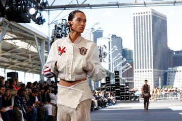 a model in a white cropped baseball jacket walks the runway in a brooklyn park with manhattan in the background