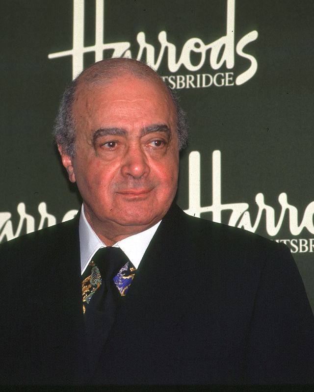 mohamed al fayed stands in front of a green harrods background, he wears a black suit jacket and tie with a multicolored collared shirt