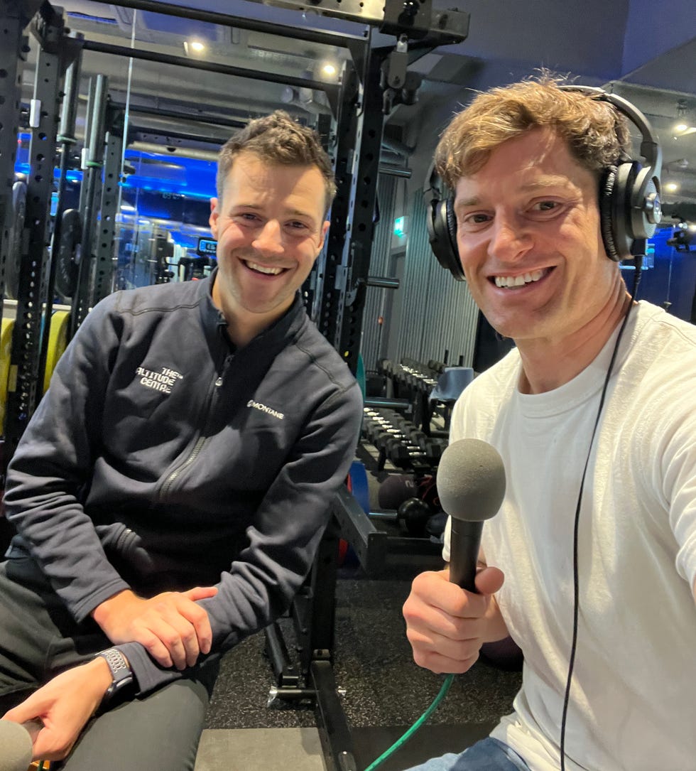 the scene captures two individuals seated in a modern gym environment one person is dressed in a dark zip up jacket while the other wears a white tshirt and holds a microphone equipment is visible in the background, indicating a workout area the lighting is bright, highlighting the gym's facilities and a contemporary atmosphere