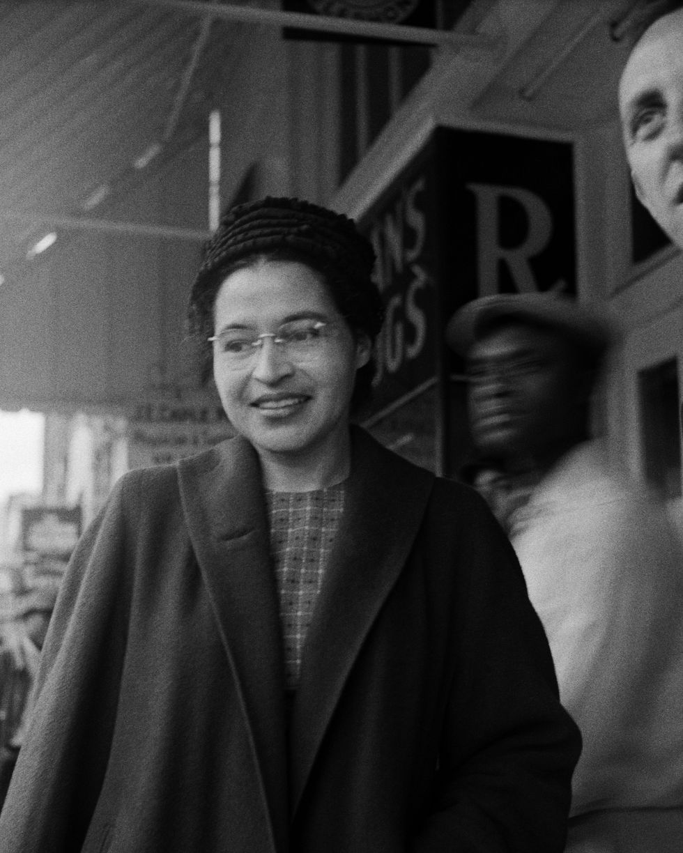 rosa parks walks outside a storefront, she wears a long coat, hat, patterned shirt, and glasses, she smiles and looks down to the left