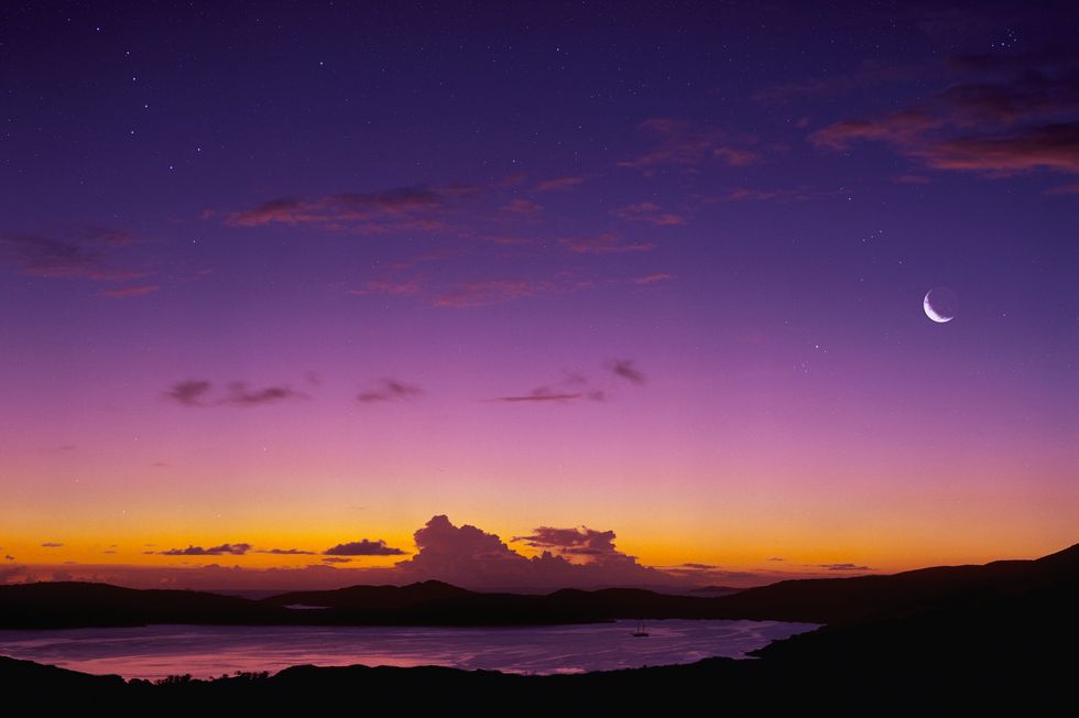 crescent moon visible at twilight in a purple and orange sky over body of water