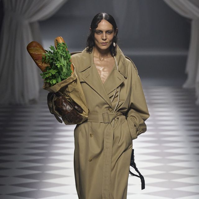 a woman in a trench coat carrying a bag of vegetables