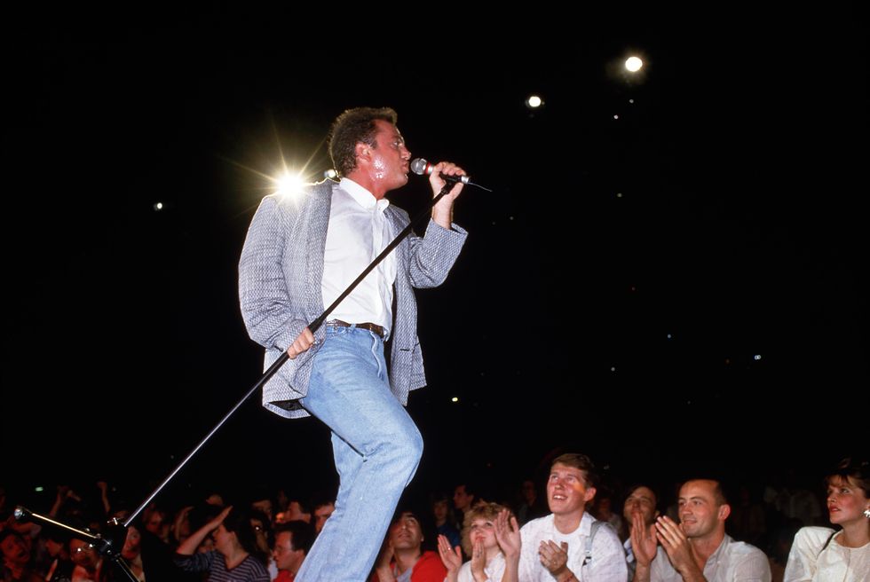 a man holds a microphone on a stand in the air as he sings to a crowd below who watch smile and clap, the performer wears a gray blazer white collared shirt and jeans