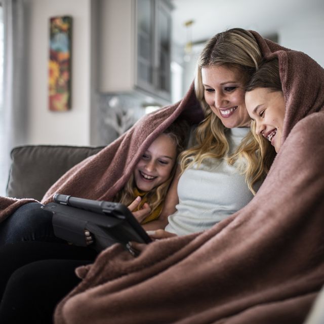 mother and tween daughters snuggled under blanket and watching a movie on digital tablet