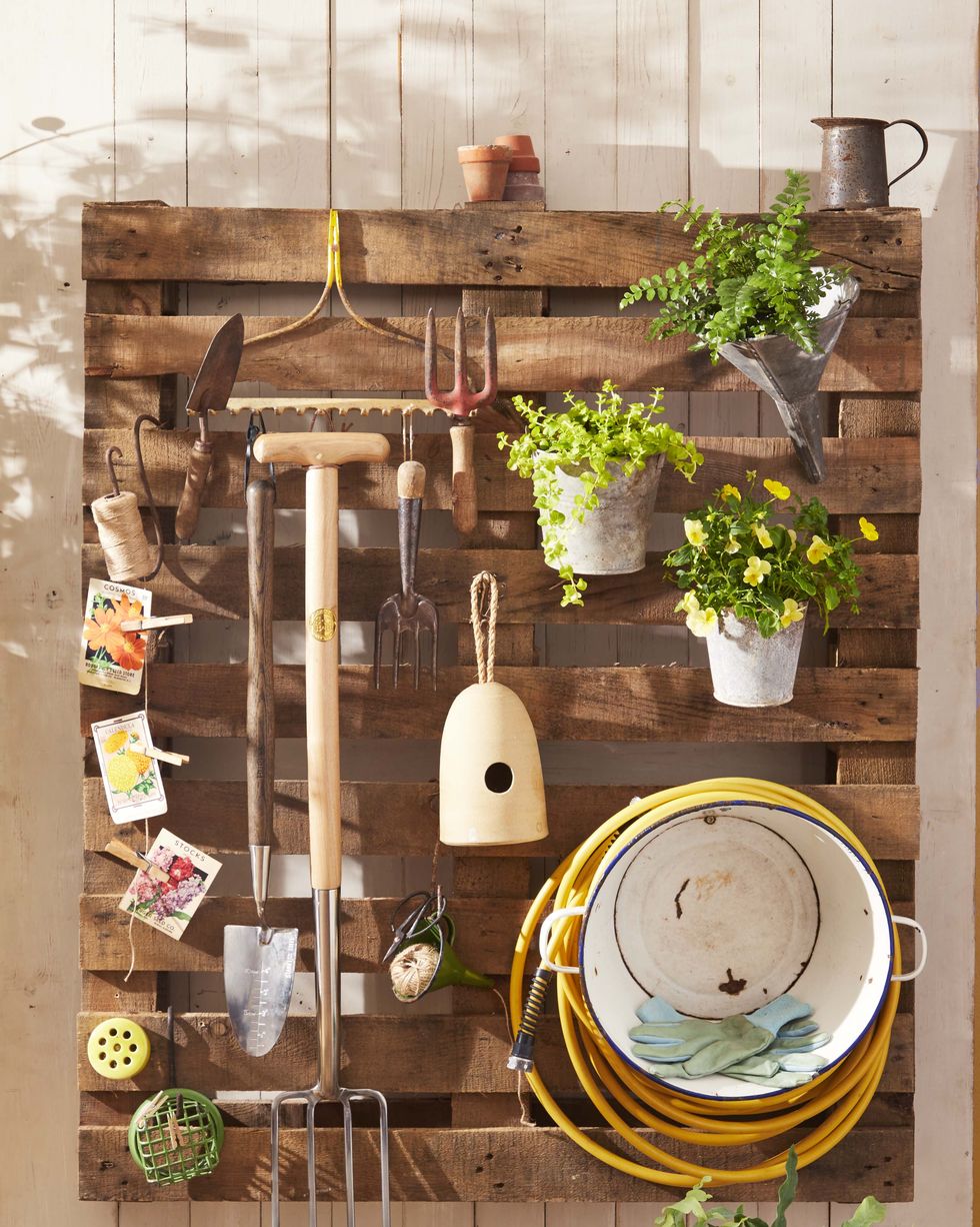 A shipping pallet hung on a white slatted wall and used to store gardening tools such as hoses and spades