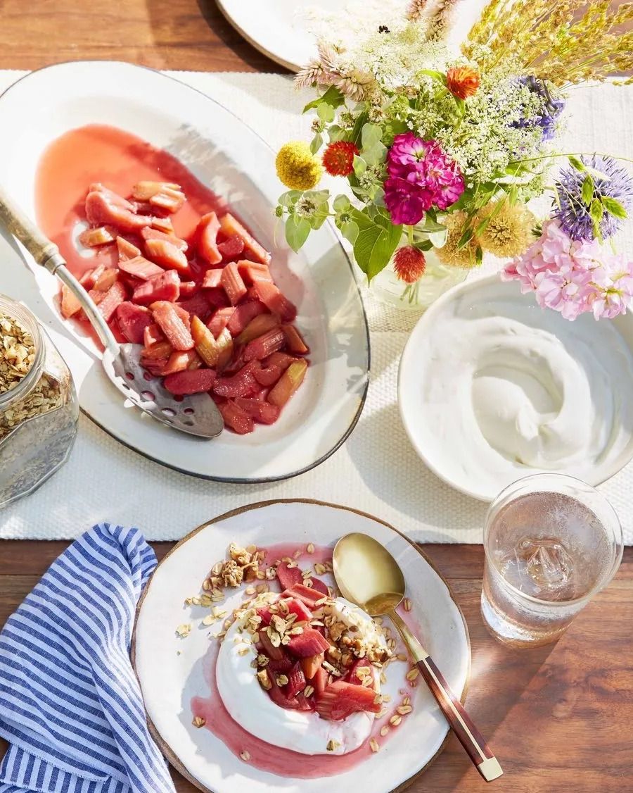 poached rhubarb with yogurt and granola on a plate with a dish of rhubarb and flowers on an outdoor table