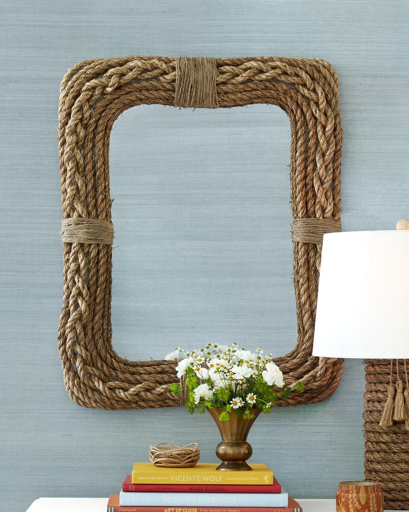 rope framed mirror hung on a gray grasscloth wall above a table that holds a lamp with a rope wrapped base, a stack of books, and a brass vase of white flowers