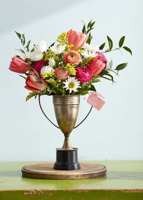 vintage metal trophy with patina filled with white daisies, pink ranunculus and tulips, and small yellow flowers, with a pink tag reading number 1 mom tied to one handle
