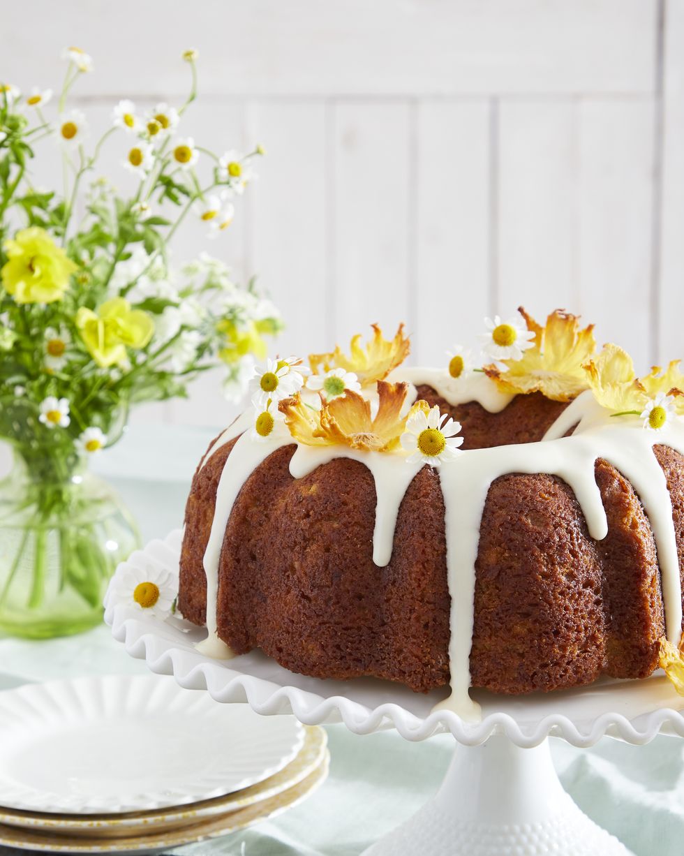 hummingbird bundt cake on a white cake stand and topped with cream cheese glaze and garnished with dried pineapple flowers and fresh camomile flowers