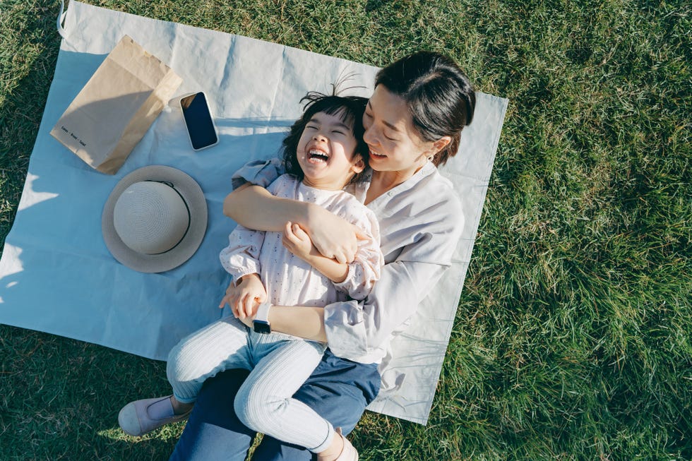 affectionate young asian mother embracing little daughter in arms, lying down on the grassy field, having fun and smiling joyfully