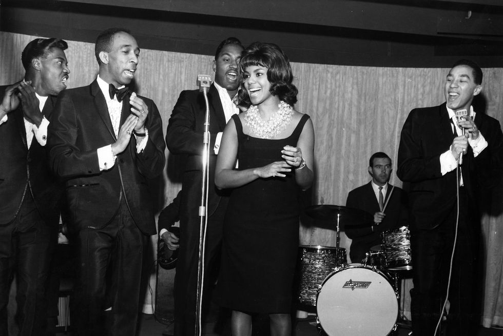 four men and a woman dance and sing on a stage as a drummer performs in the background