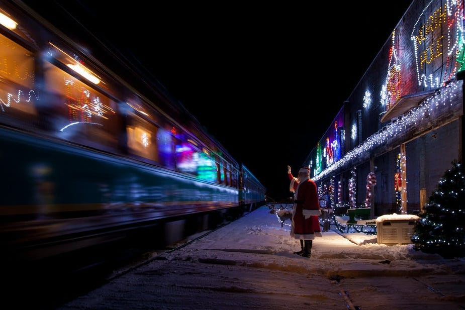 santa at a train station at night waving goodbye to a train as it pulls out of a station decorated with christmas lights and a small tree