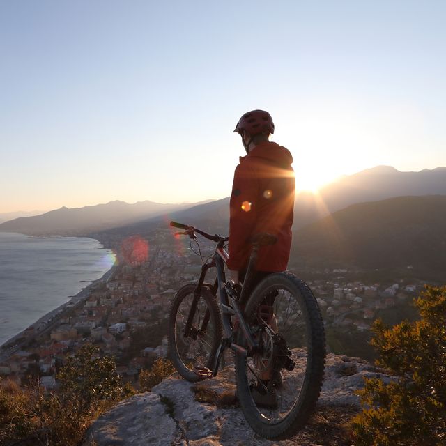 Mountain biker watches sun setting over distant mountains from hilltop