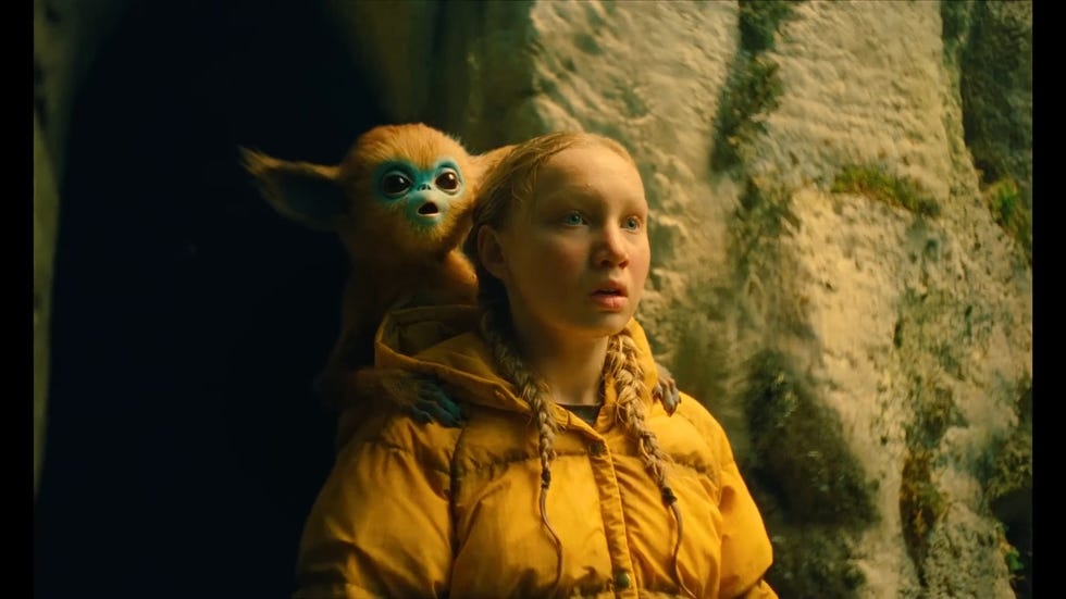a young girl with a surprised expression wears a yellow jacket with a baby ochi on her back in a scene from the legend of ochi, a good housekeeping pick for best kids' movies 2025