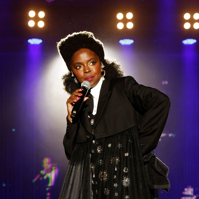 lauryn hill looks to the right as she stands on a stage and holds a microphone to her face, she wears a black and white outfit, lights shine behind her