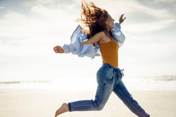 una chica con vaqueros, camisa y el pelo suelto salta en una playa