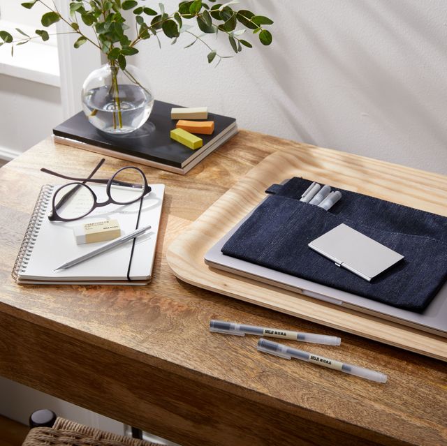 small desk with wood tray, laptop, pens, notebook, glasses and a vase with a plant set on stack of notebooks