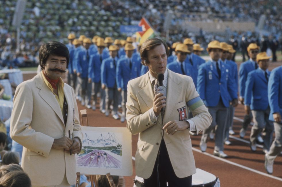 two television reporters in beige jackets looking toward the camera and speaking into microphones