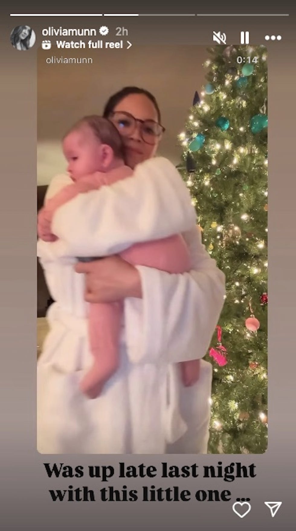 Una persona en bata de baño sosteniendo a un bebé frente a un árbol de Navidad decorado.