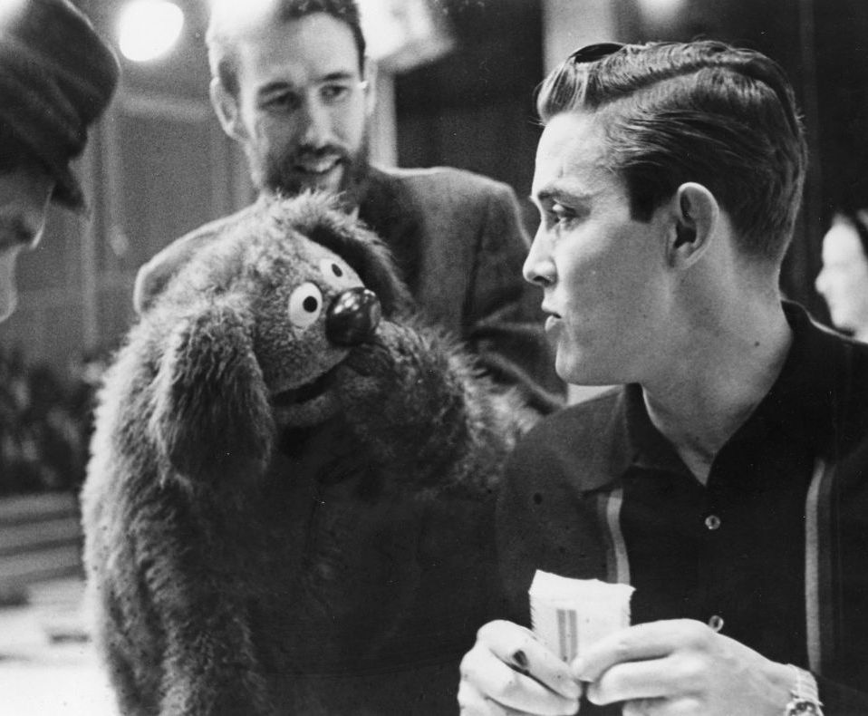 jim henson holds a muppet to jimmy deans face as the musician looks at it and holds a pack of cigarettes