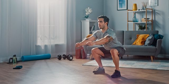 Muscular Athletic Fit Man in T-shirt and Shorts is Doing Squat Exercises at Home in His Spacious and Bright Living Room with Minimalistic Interior.