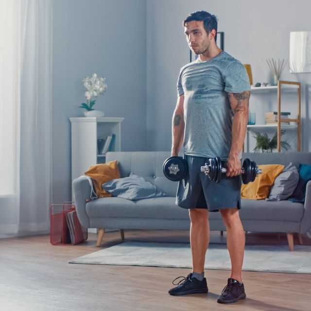 muscular athletic fit man in t shirt and shorts is doing exercises with dumbbells at home in his spacious and bright living room with minimalistic interior