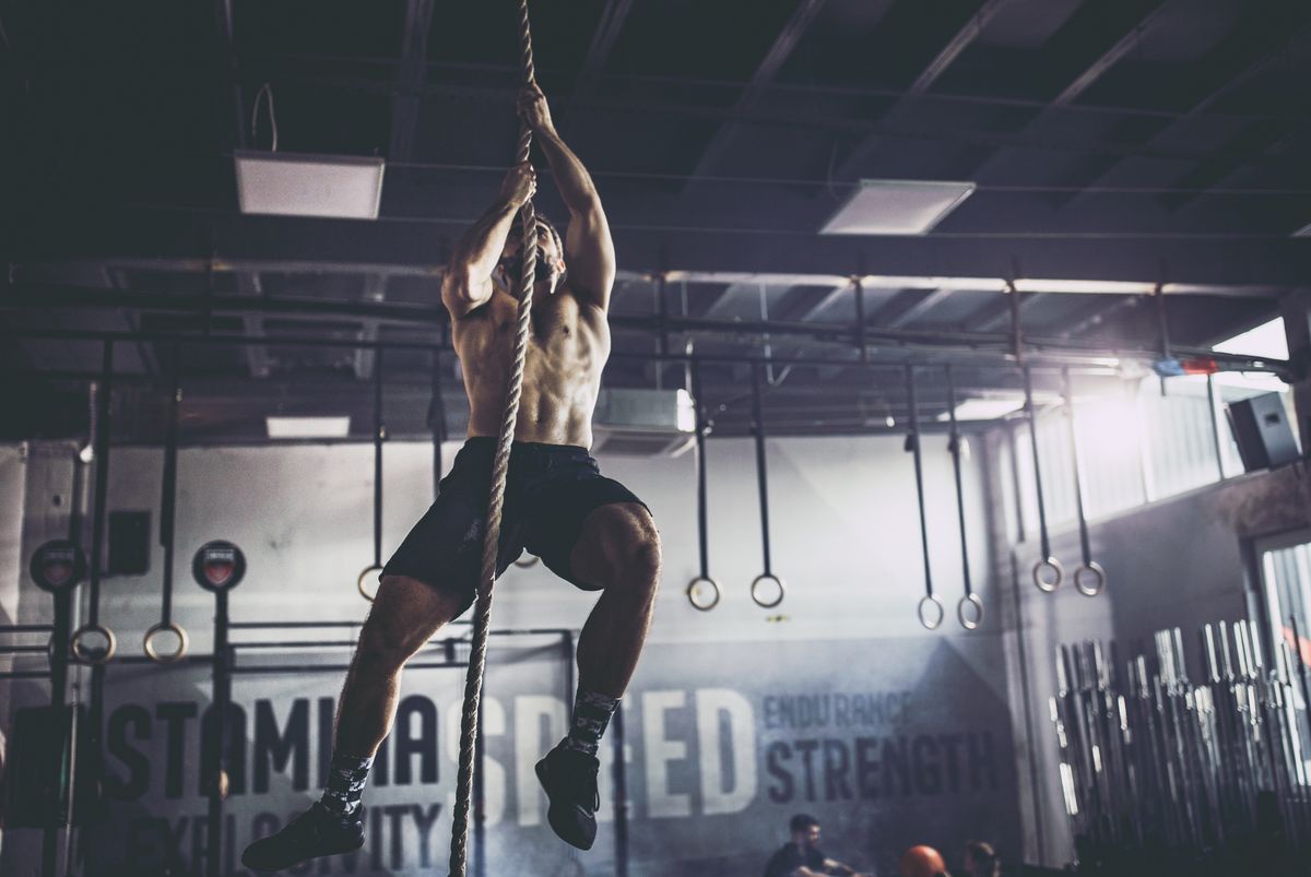 Muscular build athlete climbing up the rope in a gym.
