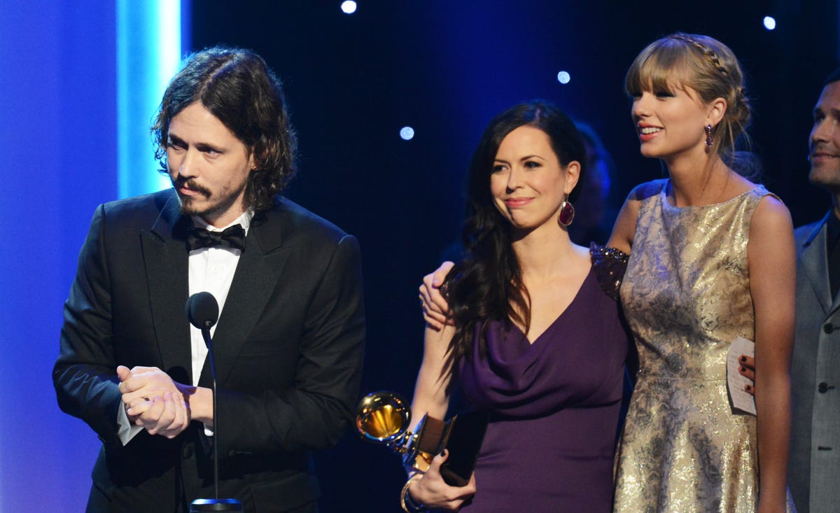 john paul white speaking while accepting a grammy award, his is wearing a black suit jacket, white collard shirt, black bowtie and his hands are clasped together, in the background to this right are joy williams and taylor swift, the women are standing and embracing while looking toward white, williams holds a grammy award