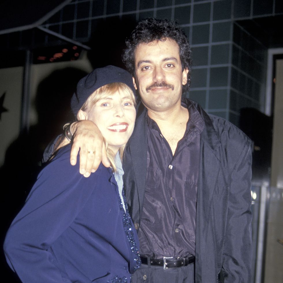 joni mitchell and larry klein smile for a photo while standing and hugging, she wears a beret and blue jacket, he wears a dark suit with a purple collared shirt