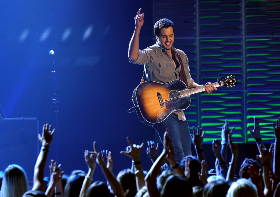 luke bryan stands on a stage above a crowd and looks down, he points one finger in the air above his head as he holds then neck of an acoustic guitar in his other hand