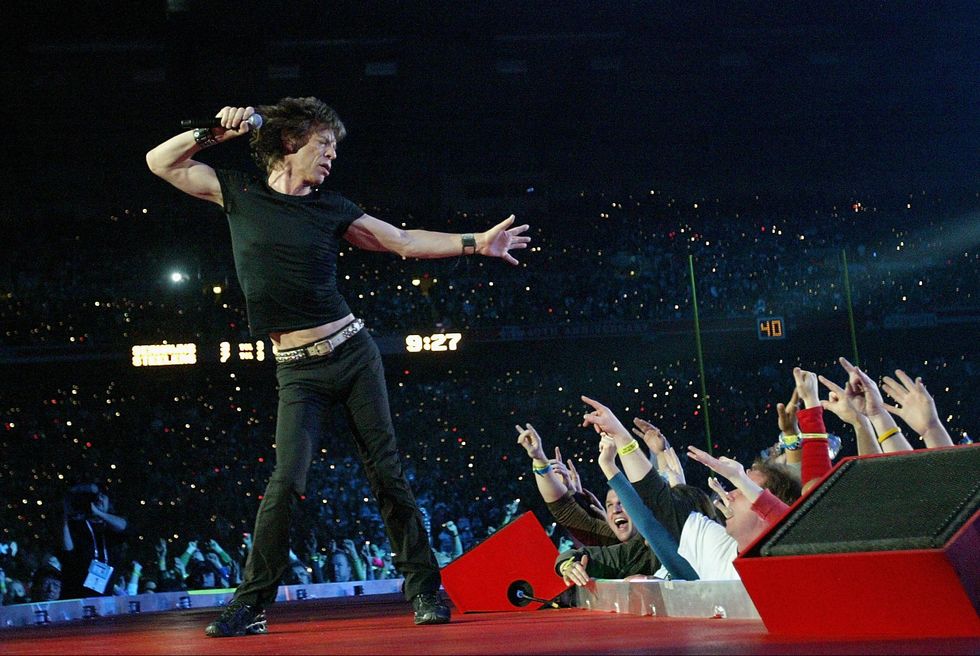 mick jagger dances on a red stage and looks down to a crowd of fans who hold up their hands, he wears a black tshirt and pants with a white belt and holds a microphone in one hand near his head