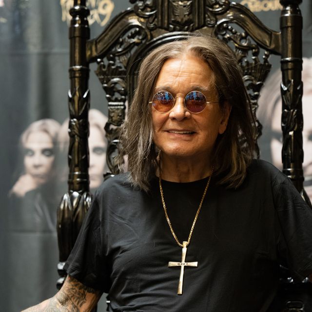 ozzy osbourne sits in an ornate high backed chair and smiles at the camera, he wears a black t shirt, large cross necklace, circular sunglasses, and a watch