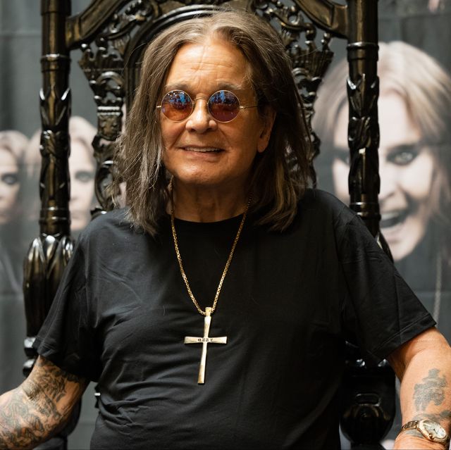 ozzy osbourne sits in an ornate high backed chair and smiles at the camera, he wears a black t shirt, large cross necklace, circular sunglasses, and a watch
