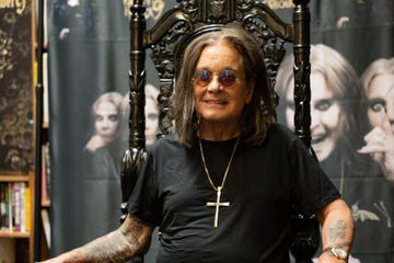 ozzy osbourne sits in an ornate high backed chair and smiles at the camera, he wears a black t shirt, large cross necklace, circular sunglasses, and a watch