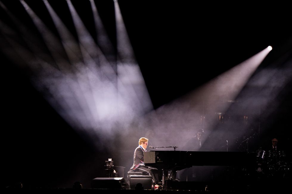 elton john plays piano and is light up by several spotlights, he wears mostly black