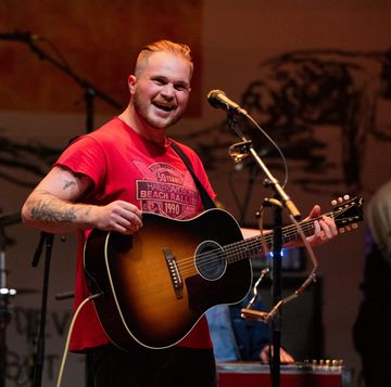 zach bryan smiling behind a microphone while playing his guitar