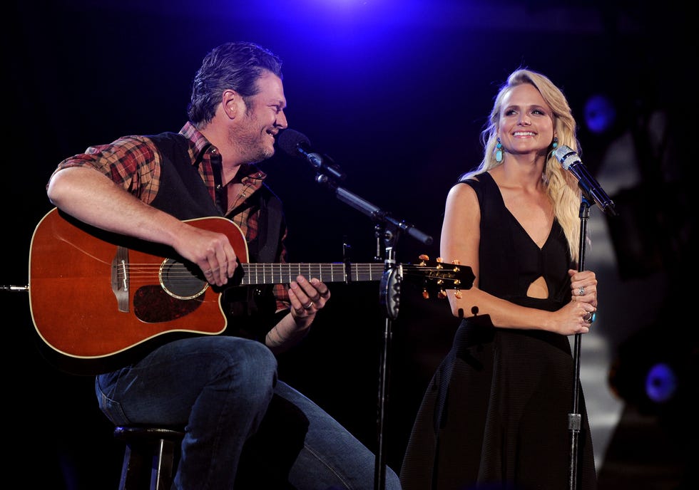 blake shelton and miranda lambert smile while on stage during a performance, he sits on a stool and plays guitar, she stands behind a microphone on a stand that she holds