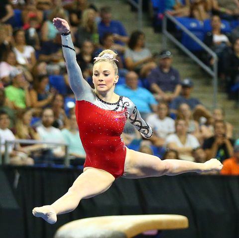 mykayla skinner leaping across the floor in a red leotard and a white and pink bow in her hair