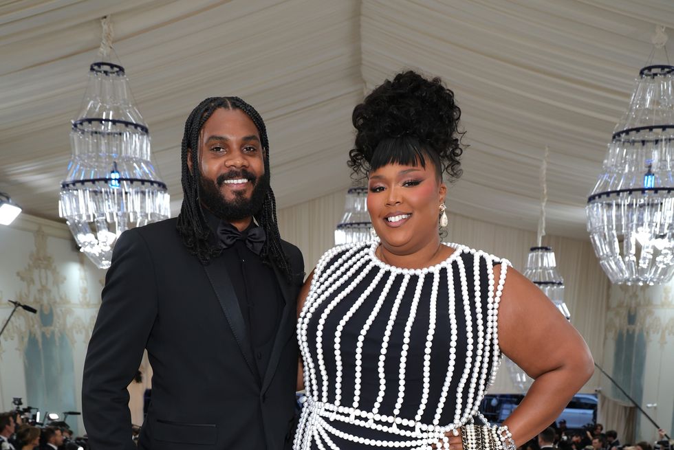 myke wright and lizzo stand next to each other embracing and smiling at the camera, he wears an all black tuxedo, she wears a black dress adorned with white pearls and pearl jewelry