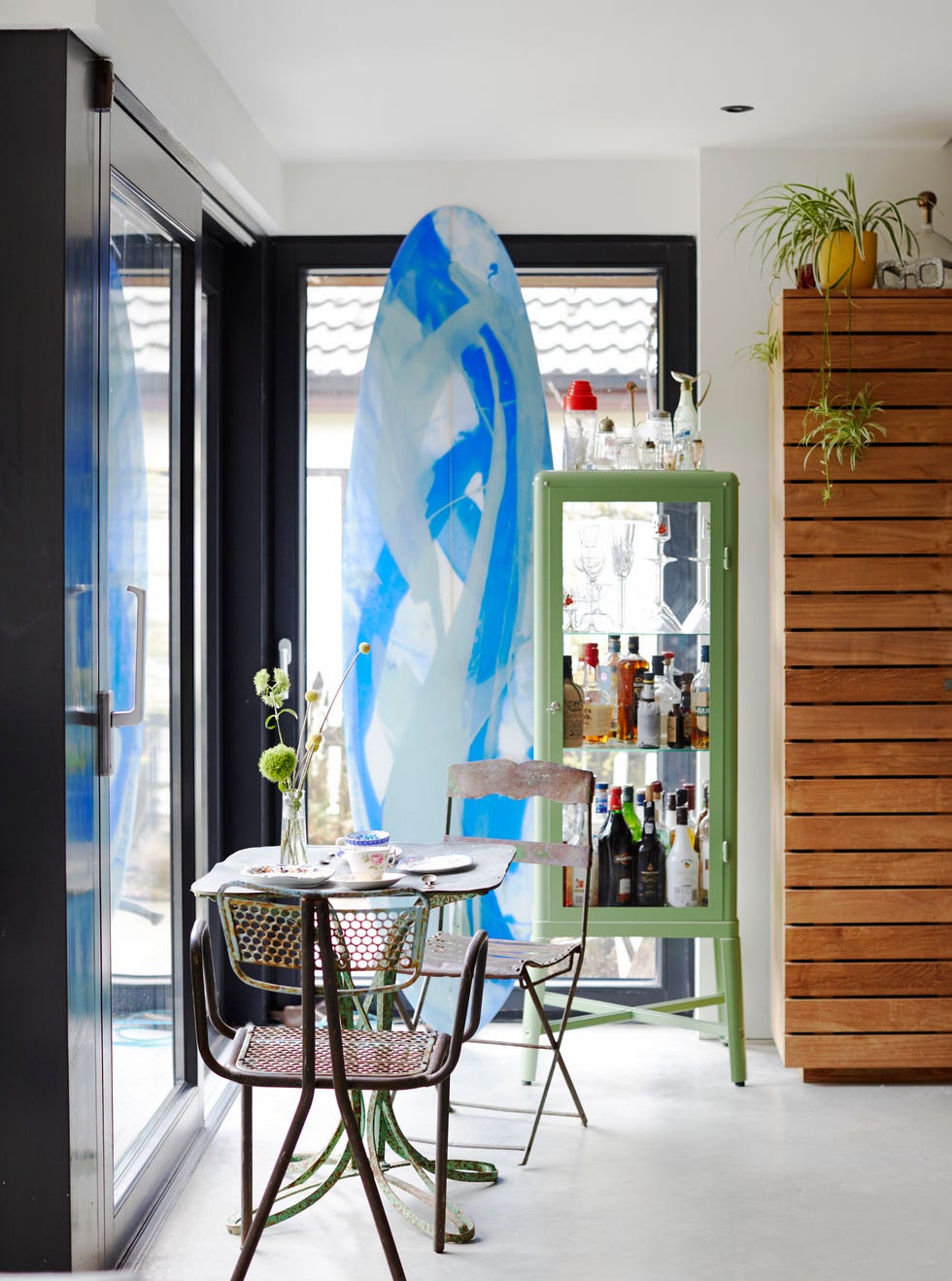 large floor to ceiling windows metal table and chairs large blue surf board and a cabinet full of bottles of alcohol