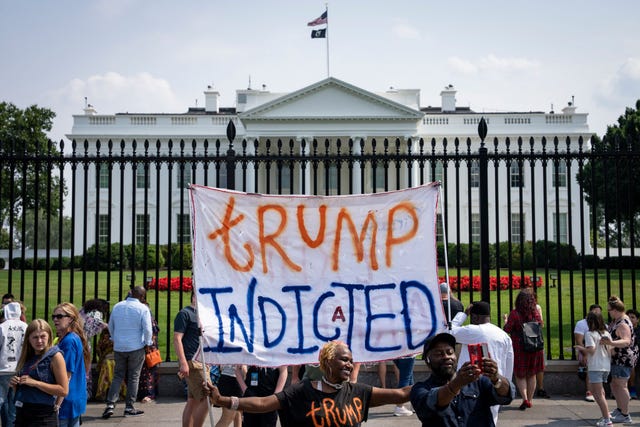 a group of protestors standing outside the gate of the white house, one of whom holds up a sign that reads trump indicted
