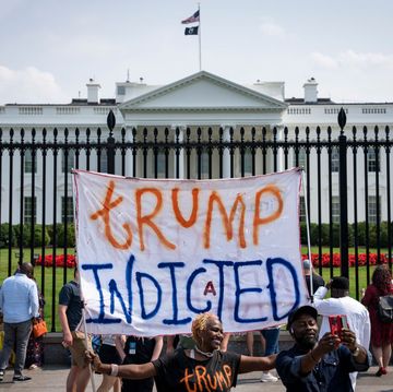 a group of protestors standing outside the gate of the white house, one of whom holds up a sign that reads trump indicted